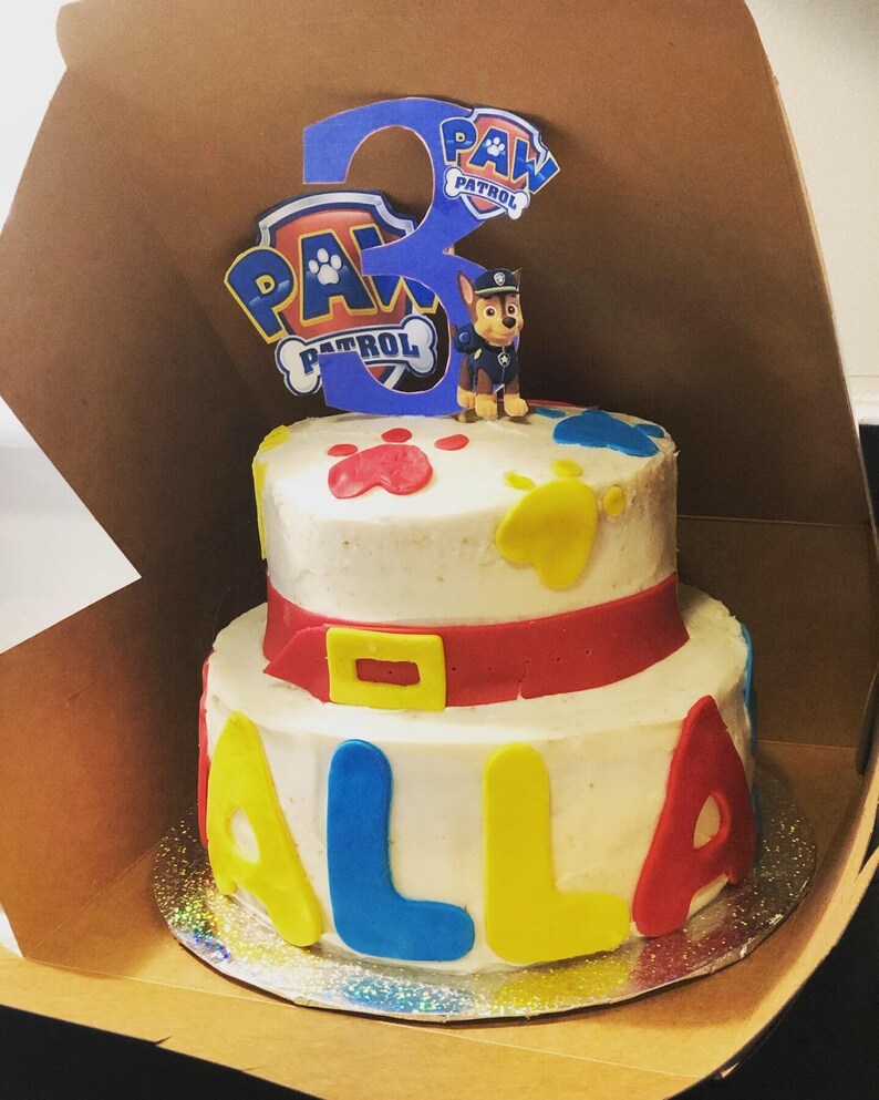 K&ouml;nnte beinhalten: Eine zweist&ouml;ckige wei&szlig;e Torte, verziert mit Pfotenabdr&uuml;cken, einem roten G&uuml;rtel und den Buchstaben "ALLA" in Blau, Gelb und Rot. Die Torte ist mit einer Paw Patrol Figur und einer blauen Zahl "3" mit den Worten "PAW PATROL" darauf verziert.