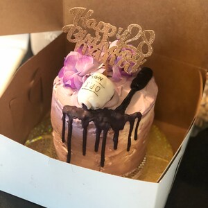 K&ouml;nnte beinhalten: Ein kleiner, einstufiger Kuchen, der mit lila Blumen, einem Schokoladen&uuml;berzug und einem "Happy 28th Birthday"-Topper verziert ist. Der Kuchen befindet sich in einer wei&szlig;en Schachtel.