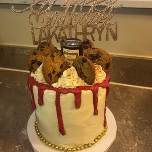 K&ouml;nnte beinhalten: Ein wei&szlig;er Kuchen mit rotem Frosting und Schokoladenkeksen darauf. Der Kuchen ist mit einem goldenen Glitzer-Topper verziert, auf dem "Happy Birthday Kathryn" steht.