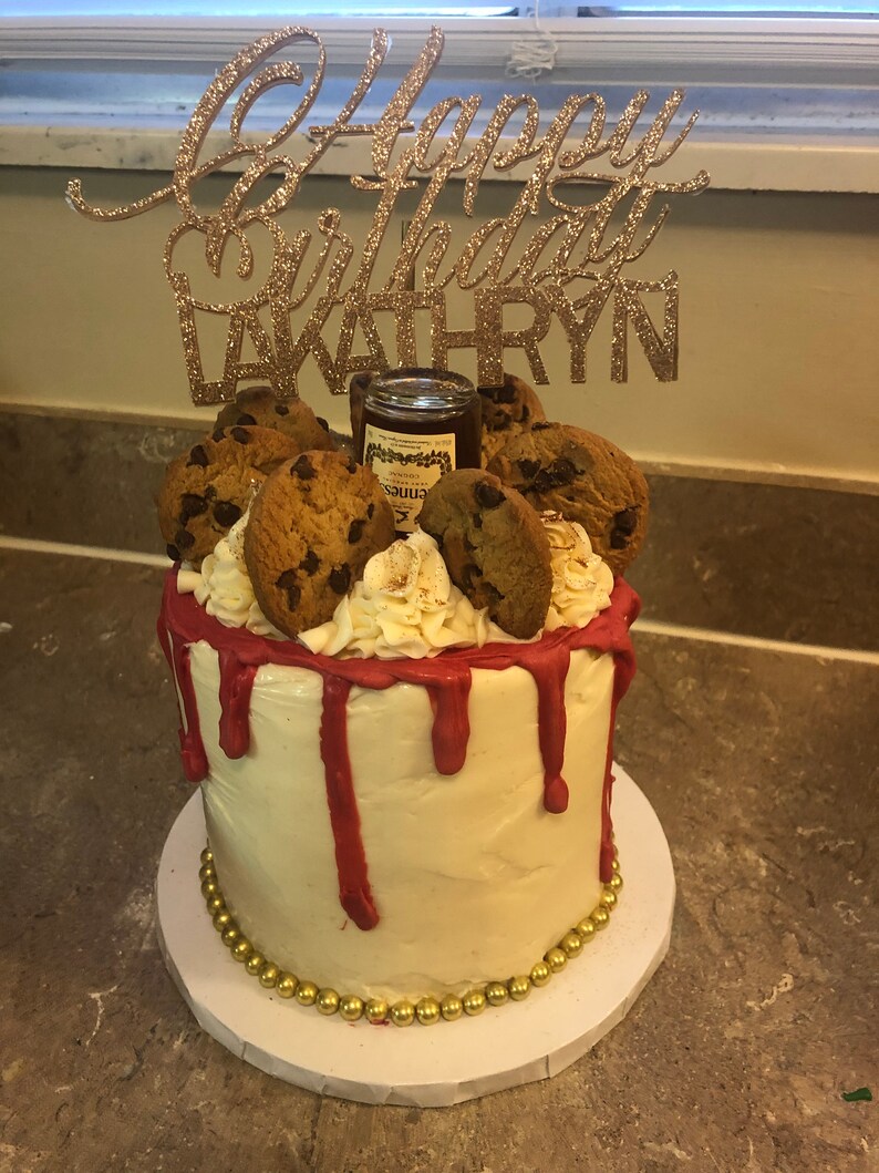 K&ouml;nnte beinhalten: Ein wei&szlig;er Kuchen mit rotem Frosting und Schokoladenkeksen darauf. Der Kuchen ist mit einem goldenen Glitzer-Topper verziert, auf dem "Happy Birthday Kathryn" steht.