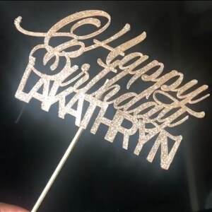 K&ouml;nnte beinhalten: Ein goldener Glitzer-Kuchentopper mit dem Text "Happy Birthday" und dem Namen "WAITHRYN" in einer kursiven Schrift.