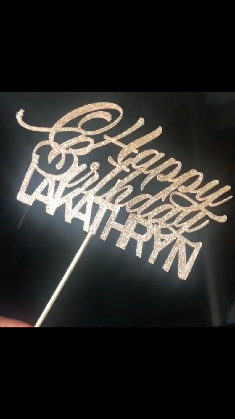 K&ouml;nnte beinhalten: Ein goldener Glitzer-Kuchentopper mit dem Text "Happy Birthday" und dem Namen "WAITHRYN" in einer kursiven Schrift.