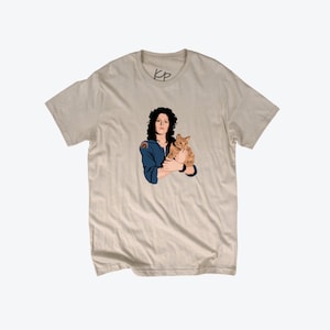 May include: A beige t-shirt with a graphic print of a woman with dark hair holding a ginger cat. The woman is wearing a blue jumpsuit and has a serious expression. The text "KP" is printed on the shirt.