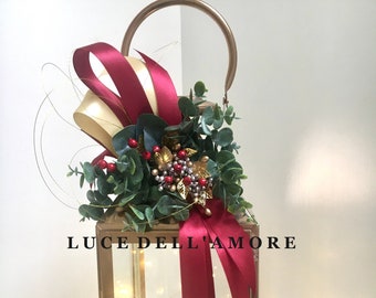 Op de afbeelding: Gouden lantaarn met een rood en gouden lint, groen en rode bessen. De lantaarn heeft een glazen voorzijde en een handvat bovenop. De tekst "LUCE DELL'AMORE" staat op de voorkant van de lantaarn.