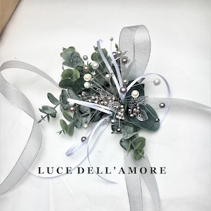 May include: A silver and white floral boutonniere with greenery, pearls, and a silver ribbon. The text "LUCE DELL'AMORE" is printed below the boutonniere.