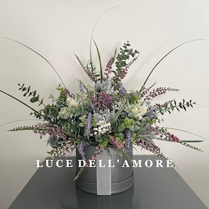 May include: A gray hatbox with a white ribbon and the text "LUCE DELL'AMORE" is filled with a bouquet of artificial flowers. The bouquet includes purple, pink, and white flowers, as well as green leaves.