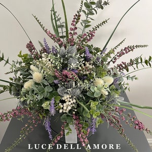 May include: A bouquet of artificial flowers in a white vase. The flowers are a mix of purple, white, and green. The vase has the text "LUCE DELL'AMORE" on the front.