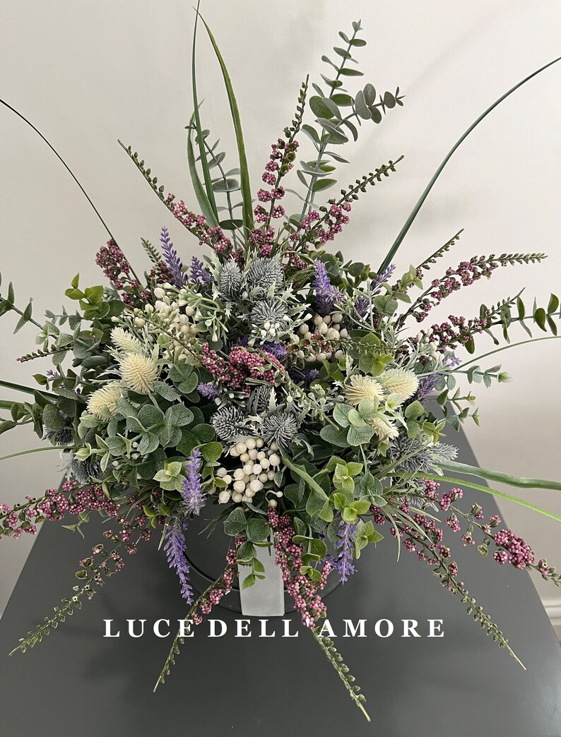 May include: A bouquet of artificial flowers in a white vase. The flowers are a mix of purple, white, and green. The vase has the text "LUCE DELL'AMORE" on the front.