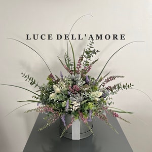 May include: A gray hat box with a white ribbon is filled with a lush arrangement of faux greenery and flowers. The flowers are a mix of white, pink, and purple. The box is labeled "LUCE DELL'AMORE".