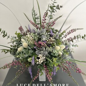 May include: A bouquet of artificial flowers in shades of purple, white, and green. The flowers are arranged in a white vase and the text "LUCE DELL'AMORE" is visible at the bottom of the image.