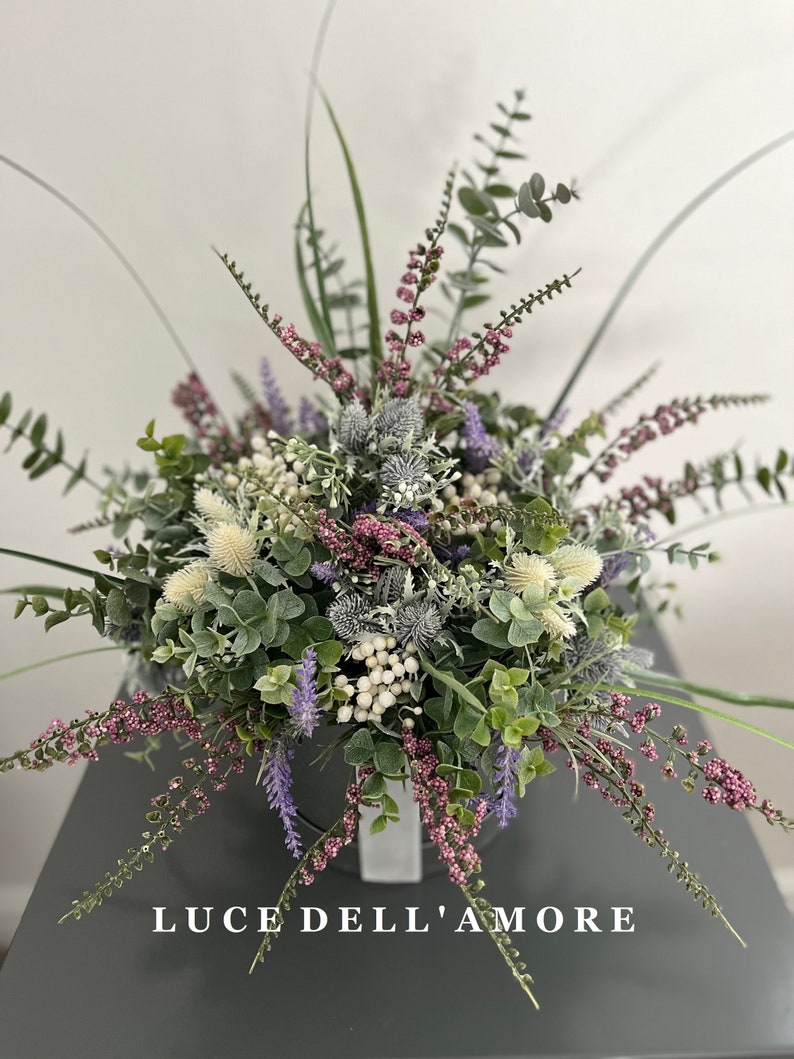 May include: A bouquet of artificial flowers in shades of purple, white, and green. The flowers are arranged in a white vase and the text "LUCE DELL'AMORE" is visible at the bottom of the image.