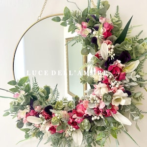 May include: A gold round mirror with a floral wreath made of pink, white, and purple flowers, greenery, and white berries. The wreath is hanging from a gold chain. The mirror reflects the text "LUCE DELL'AMORE".