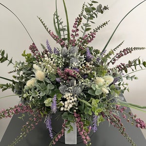 May include: A bouquet of artificial flowers in a white vase. The bouquet includes purple lavender, white berries, and green eucalyptus leaves. The vase is sitting on a gray surface. The text "LUCE DELL'AMORE" is printed on the surface.