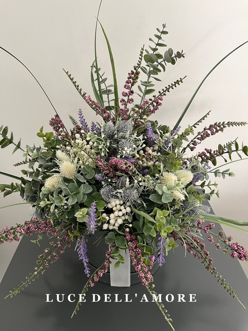 May include: A bouquet of artificial flowers in a white vase. The bouquet includes purple lavender, white berries, and green eucalyptus leaves. The vase is sitting on a gray surface. The text "LUCE DELL'AMORE" is printed on the surface.