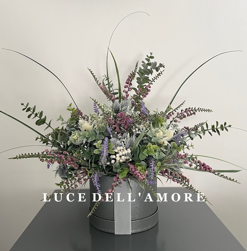 May include: A gray hatbox with a white ribbon and the text "LUCE DELL'AMORE" is filled with a bouquet of artificial flowers. The bouquet includes purple, pink, and white flowers, as well as green leaves.