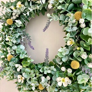 以下が含まれることがあります： 白い花と黄色の花、紫のラベンダーの枝がついた緑色のユーカリのリース。リースの下には「LUCE DELL'AMORE」という文字があります。