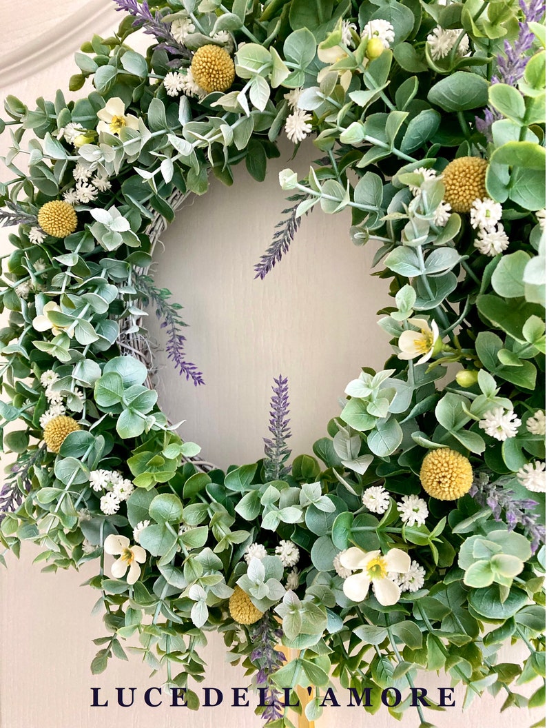 以下が含まれることがあります： 白い花と黄色の花、紫のラベンダーの枝がついた緑色のユーカリのリース。リースの下には「LUCE DELL'AMORE」という文字があります。