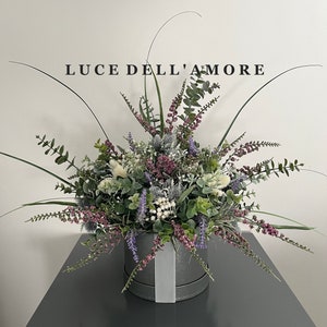 May include: A gray hatbox with a white ribbon sits on a gray surface. The hatbox is filled with a lush arrangement of faux greenery, including eucalyptus, lavender, and white berries. The text "LUCE DELL'AMORE" is printed on a white wall behind the arrangement.