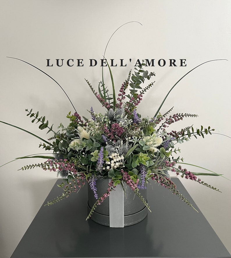 May include: A gray hatbox with a white ribbon sits on a gray surface. The hatbox is filled with a lush arrangement of faux greenery, including eucalyptus, lavender, and white berries. The text "LUCE DELL'AMORE" is printed on a white wall behind the arrangement.