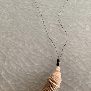 Könnte beinhalten: Eine handgefertigte Halskette mit einem Anhänger aus geschichteten, hellfarbigen Muschelteilen. Der kegelförmige Anhänger hängt an einer schwarzen Kordel, mit zwei dunklen Perlen in der Nähe der Oberseite. Die Halskette wird vor einem hellen Hintergrund präsentiert.