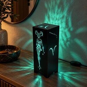 Könnte beinhalten: Eine schwarze, rechteckige Lampe mit ausgeschnittenen Motiven des Sternzeichens Widder, einem Sternbild und einer stilisierten Figur. Die Lampe strahlt hellgrünes Licht durch die Ausschnitte und wirft komplexe grüne Muster an die Wand.