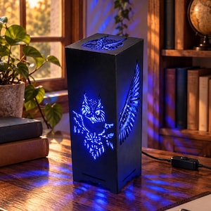 May include: A rectangular black lamp with laser-cut owl designs on the front and top, emitting a bright blue light through the cutouts. The lamp features a detailed owl figure on the front panel and a single wing design on the side panel.