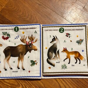 May include: A fabric book with two pages depicting woodland creatures. One page shows a moose with text "Can you count 1 woodland animal?" The other page shows a wolf and fox with text "Can you count 2 woodland friends?"