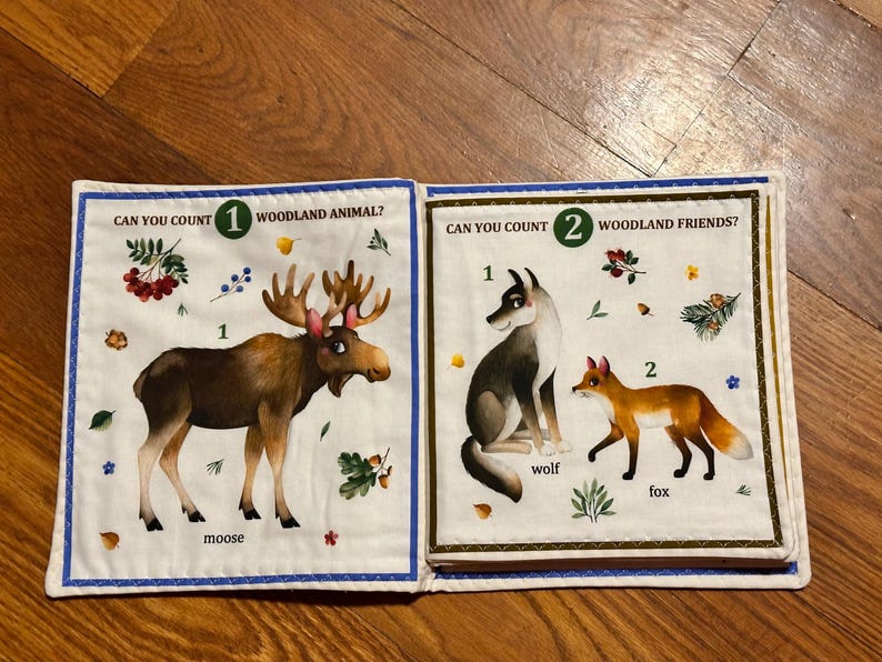 May include: A fabric book with two pages depicting woodland creatures. One page shows a moose with text "Can you count 1 woodland animal?" The other page shows a wolf and fox with text "Can you count 2 woodland friends?"