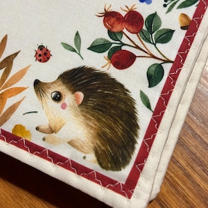 May include: A white fabric item featuring a charming hedgehog illustration, with floral and berry details. A red border with white stitching frames the design. The image also includes a ladybug and text.