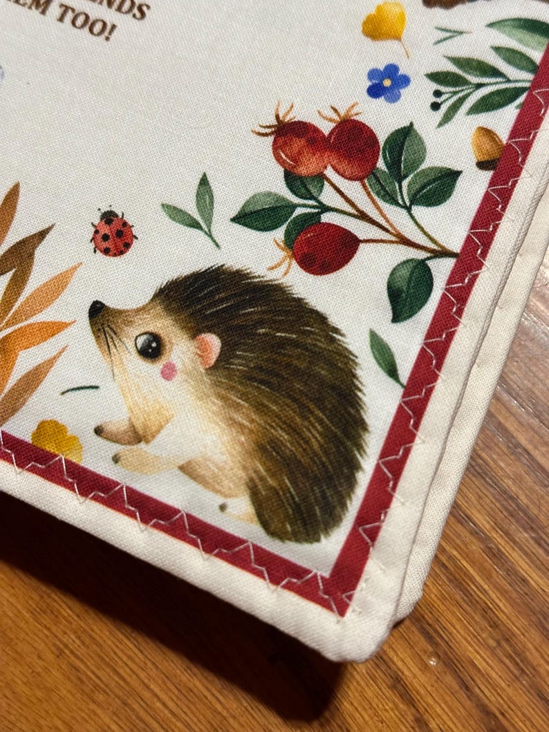 May include: A white fabric item featuring a charming hedgehog illustration, with floral and berry details. A red border with white stitching frames the design. The image also includes a ladybug and text.
