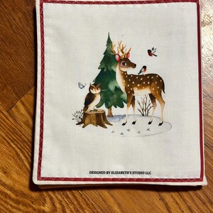 May include: Square white fabric with a red border. It depicts a woodland scene with a spotted deer, green tree, owl, and birds. The text "DESIGNED BY ELIZABETH'S STUDIO LLC" is printed at the bottom. A charming, decorative item.