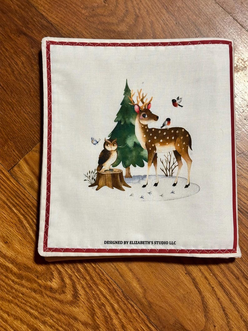 May include: Square white fabric with a red border. It depicts a woodland scene with a spotted deer, green tree, owl, and birds. The text "DESIGNED BY ELIZABETH'S STUDIO LLC" is printed at the bottom. A charming, decorative item.