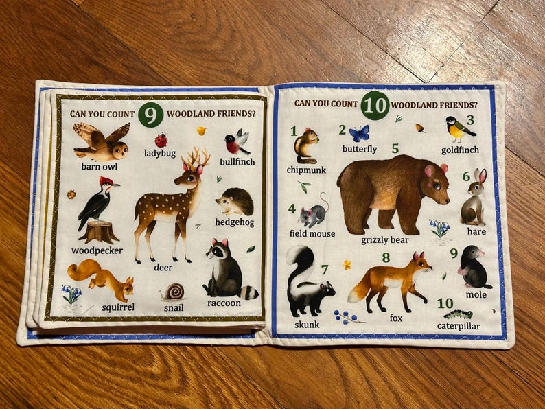 May include: A children's fabric book featuring illustrations of woodland creatures. The pages show numbers and animal names, including "barn owl," "deer," and "grizzly bear." Text reads "Can You Count 9 Woodland Friends?" and "Can You Count 10 Woodland Friends?"