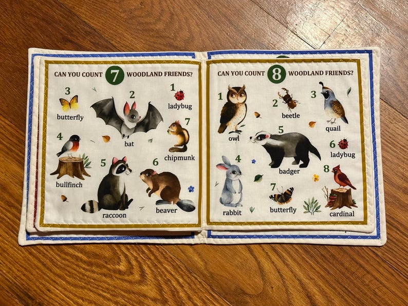 May include: A children's fabric book open to two pages, featuring illustrations of woodland creatures. The left page shows a butterfly, bat, raccoon, and chipmunk. The right page shows an owl, beetle, badger, and rabbit. Text reads "Can You Count Woodland Friends?"