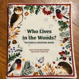 May include: A children's book cover with the title "Who Lives in the Woods?" surrounded by illustrations of woodland animals and foliage. The cover includes the text "Picture & Counting Book" and is framed with a red stitched border.