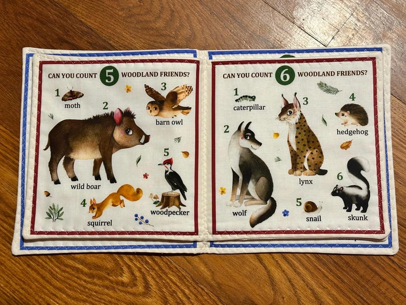 May include: A fabric book featuring woodland creatures. The left page displays a moth, wild boar, barn owl, squirrel, and woodpecker. The right page shows a caterpillar, wolf, lynx, hedgehog, snail, and skunk. Text reads "CAN YOU COUNT WOODLAND FRIENDS?"