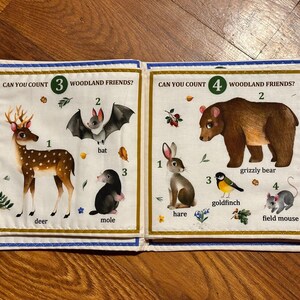 May include: A fabric book open to two pages with woodland animal illustrations. One page shows a deer, bat, and mole. The other page shows a hare, grizzly bear, goldfinch, and field mouse. Text reads "Can you count woodland friends?"