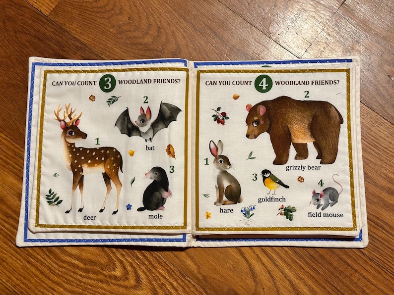 May include: A fabric book open to two pages with woodland animal illustrations. One page shows a deer, bat, and mole. The other page shows a hare, grizzly bear, goldfinch, and field mouse. Text reads "Can you count woodland friends?"
