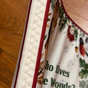 May include: Close-up of a fabric item with a cream background, red stripes, and a decorative stitched edge. The fabric features a forest-themed print with text that reads "Who lives in the Woods? & Counting Book."