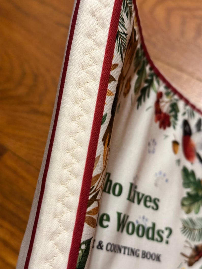 May include: Close-up of a fabric item with a cream background, red stripes, and a decorative stitched edge. The fabric features a forest-themed print with text that reads "Who lives in the Woods? & Counting Book."