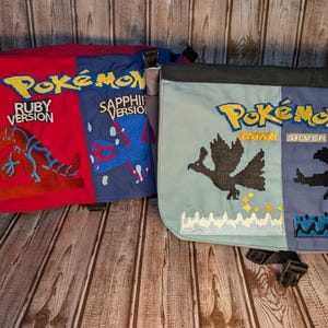 May include: Two messenger bags with the Pokémon logo. One bag is red and blue with "Ruby Version" and "Sapphire Version" text. The other bag is light blue and blue with "Gold" and "Silver" text. Each bag has a black strap and buckle.
