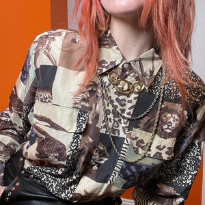 May include: A patterned button-down shirt with animal print squares in brown, black, and cream. The shirt has two chest pockets and is accessorized with a gold-toned necklace.