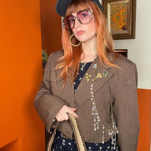 Könnte beinhalten: Ein brauner Blazer mit einer dekorativen Kette aus klaren Perlen und goldfarbenen Akzenten. Der Blazer wird mit einem marineblauen Kleid mit Blumenmuster, einer beigefarbenen Handtasche und einer großen rosa getönten Sonnenbrille kombiniert. Eine dunkelblaue Baseballkappe und goldene Creolen vervollständigen den Look.