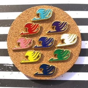 Puede incluir: Un conjunto de diez pines de esmalte con el logotipo de Fairy Tail en varios colores, incluyendo azul, rojo, rosa, blanco, verde, amarillo y dorado. Los pines se muestran en un tablero de corcho.
