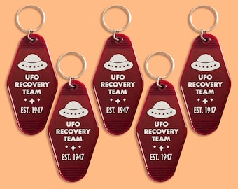 Könnte beinhalten: Fünf rote Schlüsselanhänger in Form von Motel-Schlüsseln mit dem Text "UFO Recovery Team + EST. 1947" und einer weißen Silhouette eines UFOs.