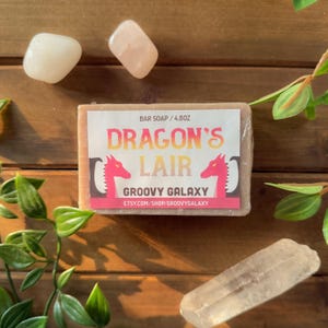May include: A bar of soap with the text "DRAGON'S LAIR" and "GROOVY GALAXY" on a label. The soap is 4.8oz. Two pink dragon illustrations flank the text. The image also includes clear and pink crystals and green foliage.