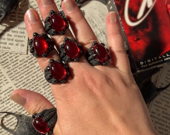 May include: Six dark gray rings with large, oval, red gemstones are displayed on a hand. The rings have a textured, claw-like design around the stones. The background includes a vintage movie case and newspaper clippings.