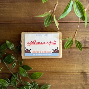May include: A rectangular bar soap with a label that reads "Mothman Mall GROOVY GALAXY" and "BAR SOAP / 4.8OZ". The soap is light brown and wrapped in clear plastic. Green leaves and stems are in the background.