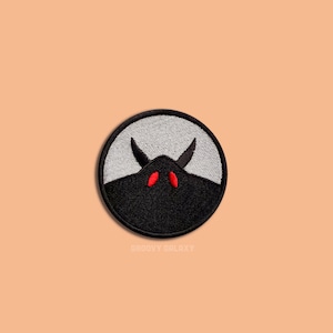 May include: A black and white embroidered patch with a black silhouette of a creature with red eyes and horns. The patch is circular and has a white background.
