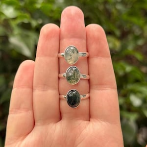 May include: Three silver rings with oval-shaped gemstones. The top ring has a light green stone, the middle ring has a green and brown stone, and the bottom ring has a dark green stone. The rings are displayed on a hand.